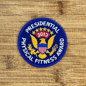 Könnte beinhalten: Ein kreisförmiger blauer Presidential Physical Fitness Award-Aufnäher. Der Aufnäher zeigt einen goldenen Adler mit ausgebreiteten Flügeln, einen rot-weiß gestreiften Schild und das Jahr 2013. Der Text "Presidential Physical Fitness Award" ist in Weiß.