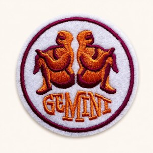 May include: A circular embroidered patch featuring a white background with a dark red border. The design displays two stylized orange figures facing away from each other, positioned above the orange embroidered text that reads GEMINI.