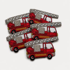 Loose pile of red embroidered fire truck patches with silver ladders, yellow accents, black wheels, and detailed stitched texture on a clean white background.