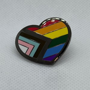 May include: A rainbow heart-shaped pin with a black outline. The rainbow is made up of red, orange, yellow, green, blue, indigo, and violet stripes. The pin is a geometric design with a black and white pattern in the center.