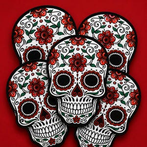 Loose pile of Day of the Dead sugar skull embroidered patches with red flowers, green leaves, black scrollwork, and bold black borders on a deep red square background.