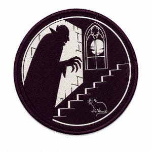 May include: A circular embroidered patch featuring a dark purple and white graphic of a caped figure with clawed hands standing on a staircase. A small rat sits at the base of the stairs, and a gothic arched window with a full moon is visible.