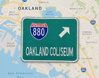 May include: A rectangular embroidered patch featuring a green background with a white border. It displays a blue and red Interstate 880 shield, a white directional arrow, and the white text OAKLAND COLISEUM printed in capital letters.