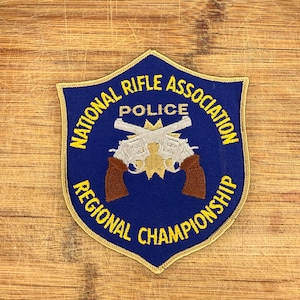 May include: A shield-shaped embroidered patch with a dark blue background and a gold border. It features two crossed silver revolvers with brown grips, a gold star emblem, and yellow text reading National Rifle Association Police Regional Championship.