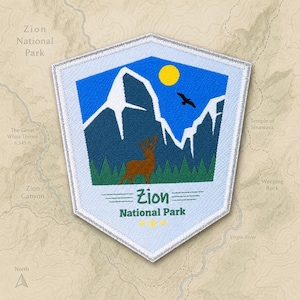 Peut inclure: Un écusson brodé en forme de bouclier représentant un paysage de montagnes bleu foncé aux sommets enneigés, un soleil jaune, un oiseau noir et un cerf brun dans une forêt verte. Le texte indique Zion National Park avec trois étoiles jaunes.