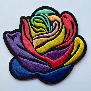 May include: A rainbow-colored embroidered rose patch. The rose is made of red, orange, yellow, green, blue, and purple thread. The patch is outlined in black thread.