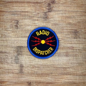 May include: A circular embroidered patch with a black background and a blue border. The text RADIO DISPATCHER is stitched in yellow. In the center, a yellow circle is surrounded by red lightning bolt graphics radiating outward in four directions.
