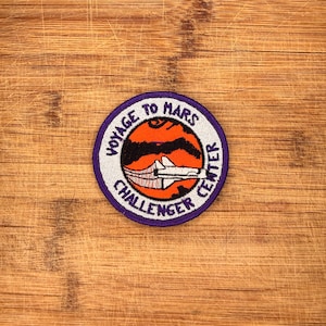 May include: A circular embroidered patch featuring a white border with the purple text VOYAGE TO MARS CHALLENGER CENTER. The center depicts an orange planet with black surface details and a white space shuttle illustration flying across it.
