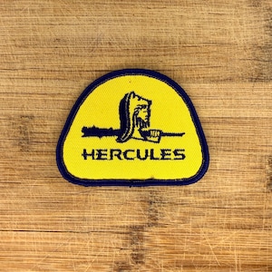 May include: A yellow patch with a blue border and the word "HERCULES" in black text. The patch features a blue silhouette of a classical bust and a horizontal line.