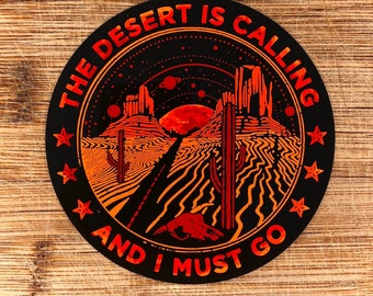 May include: A circular sticker featuring a black background with orange and red desert landscape graphics, including cacti, rock formations, a road, and planets. The text reads THE DESERT IS CALLING AND I MUST GO, framed by small stars.