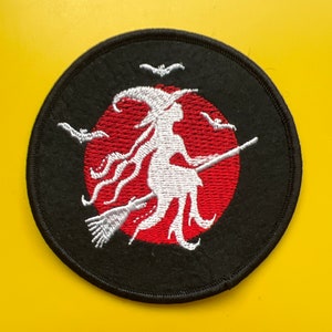 Peut inclure: Un écusson brodé noir et rouge représentant une silhouette blanche de sorcière volant sur un balai devant un soleil rouge. Trois chauves-souris blanches volent au-dessus de la sorcière.