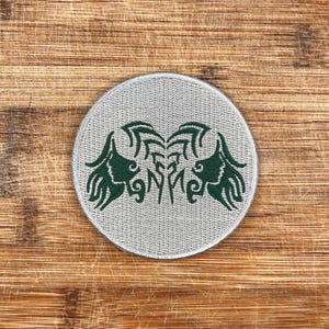 Puede incluir: Un parche redondo bordado de color gris con un diseño estilizado verde de dos pájaros enfrentados con las alas extendidas.