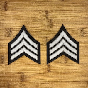 以下が含まれることがあります： 木製の背景に、2つの黒と銀のシェブロンが表示されています。これらは軍の階級章である可能性があります。各シェブロンは、黒い縁取りの中に3つの銀色のストライプがあり、対称的で上向きのデザインになっています。これらのパッチは、制服への適用を目的としています。