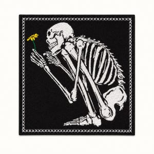 May include: A square embroidered patch featuring a white skeleton in a crouching position on a black background. The skeleton holds a small yellow flower with a green stem. The border of the patch is decorated with a white cross-stitch pattern.
