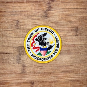 Op de afbeelding: Een ronde geborduurde patch met een zwarte adelaar, een rood lint en een schild met rode en witte strepen. De gele rand bevat de zwarte tekst THE TOWN OF CICERO INCORPORATED FEB. 26, 1967, rondom een centrale afbeelding van een zon.