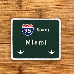 May include: Green and white interstate highway sign with a white border. The sign reads "Interstate 95 South Miami" with two white downward arrows.