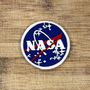 May include: A circular embroidered patch featuring a dark blue background with the white text NASA in the center. A red curved line crosses the letters, and white star-like shapes are scattered across the blue field with a white border.