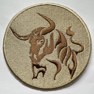 Peut inclure: Un patch brodé beige avec une silhouette de taureau brune. La tête du taureau est tournée vers le côté gauche du patch. Les cornes du taureau sont courbées et pointues. La queue du taureau est enroulée.
