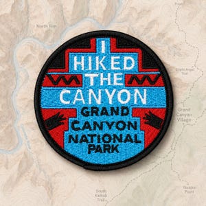 May include: A circular embroidered patch with a black border. The design features a stepped geometric pattern in bright blue and red. The text reads I HIKED THE CANYON GRAND CANYON NATIONAL PARK in white and black lettering against the blue background.