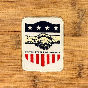 Könnte beinhalten: Ein rechteckiger Aufnäher mit cremefarbenem Rand. Der obere Teil zeigt drei weiße Sterne auf marineblauem Hintergrund. Darunter sind zwei Hände im Handschlag verschränkt. Der untere Teil hat rote vertikale Streifen mit dem Text "UNITED STATES OF AMERICA".