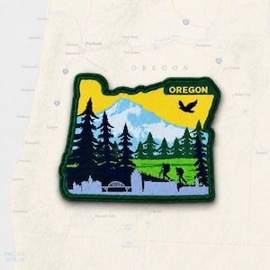 May include: A shaped embroidered patch in the outline of the state of Oregon. The design features a yellow sky, a light blue mountain, dark green pine trees, and a light green meadow. Two hikers and a bird are visible with the text OREGON.
