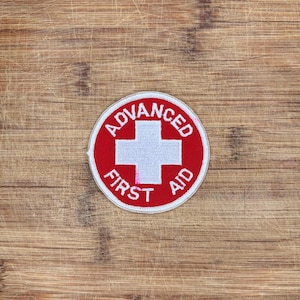 Może przedstawiać: Okrągła haftowana naszywka z czerwonym tłem i białą obwódką. W centrum znajduje się biały symbol krzyża. Napis ADVANCED FIRST AID jest wyhaftowany białymi wielkimi literami, układającymi się w łuk wokół górnej i dolnej części krzyża.