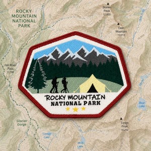 May include: A hexagonal embroidered patch featuring a landscape of snow-capped mountains, a dark green forest, a yellow tent, and two hikers. The text reads ROCKY MOUNTAIN NATIONAL PARK with three yellow stars below. The patch has a red border.