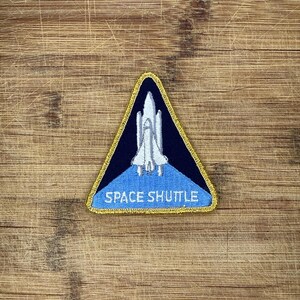 Può includere: Una toppa ricamata triangolare che raffigura una navetta spaziale bianca su sfondo blu scuro. La parte inferiore della toppa è azzurra con la scritta bianca SPACE SHUTTLE. L'intera toppa è rifinita con un bordo dorato.