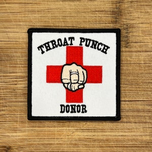 May include: White embroidered patch with a red cross and a fist in the centre. The text "Throat Punch Donor" is above and below the cross.