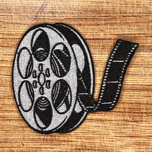 May include: A black and white embroidered patch of a film reel with a strip of film attached. The film reel has a silver colour and the film strip is black.