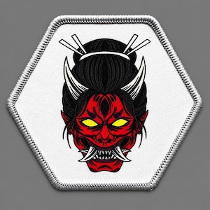 May include: A hexagonal embroidered patch featuring a red demon face with yellow eyes, sharp white fangs, and black hair styled in a bun with two white hair sticks. The patch has a white background and a silver-colored stitched border edge.
