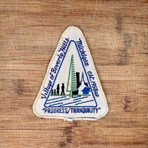 May include: A triangular patch with a cream-colored border and a design featuring the text "Village of Beverly Hills Michigan est. 1958AD". The patch includes a blue and green illustration of a tree, people, and buildings. The text "PROGRESS/TRANQUILITY" is also present.