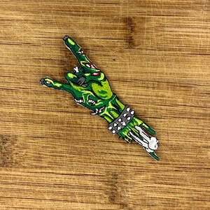 Zombie hand throwing devil horns—green embroidered iron-on patch with bone and stud cuff—shown horizontal on wood for scale.