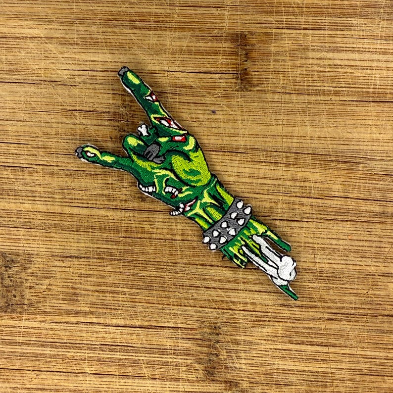 Zombie hand throwing devil horns—green embroidered iron-on patch with bone and stud cuff—shown horizontal on wood for scale.