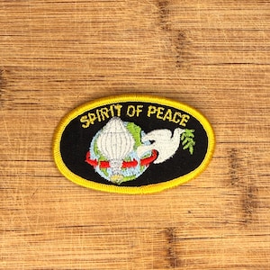 May include: An oval embroidered patch with a black background and a yellow border. It features a globe, a white parachute, and a white dove holding a green olive branch. The text SPIRIT OF PEACE is embroidered in yellow letters at the top.