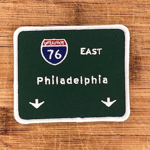 May include: Green and white embroidered patch with a highway sign that reads "Interstate 76 East Philadelphia" with two white arrows pointing down.