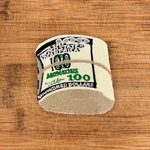 Puede incluir: Un calcetín enrollado con un diseño que se asemeja a un billete de 100 $. El calcetín presenta el texto "UNITED STATES OF AMERICA", "100" y "ONE HUNDRED DOLLARS" en negro, verde y blanco. Una banda elástica de color canela envuelve el centro.
