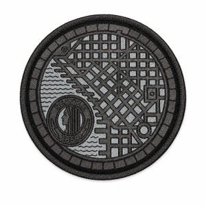 May include: A circular embroidered patch featuring a gray and black manhole cover design. The pattern includes a grid, wavy lines, and a circular emblem with a profile silhouette and the text SEATTLE 1869. The patch has a thick black stitched border.