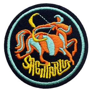 May include: A circular embroidered patch featuring a dark navy background with a light blue border. The design depicts an orange and light blue centaur archer holding a yellow bow. The word SAGITTARIUS is embroidered in yellow at the bottom.