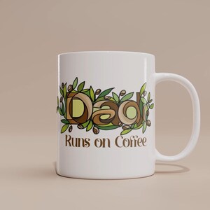Op de afbeelding: Witte keramische mok met het woord "Dad" in een bruin en groen bloemenontwerp. De tekst "Runs on Coffee" staat onder het woord "Dad" afgedrukt.