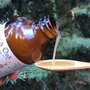 May include: A brown glass bottle with a white label reading & Oak, pouring a light-colored viscous liquid into a wooden spoon. The label includes text describing the product as containing myrrh and other ingredients in a clear, readable font.