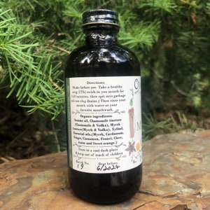 May include: A dark amber glass bottle with a black screw cap, containing an 8 oz liquid product. The white label features text including directions for use, organic ingredients like sesame oil and myrrh, batch number 19, and a best before date of 6/2024.