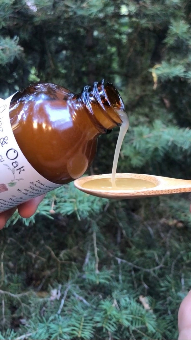 May include: A brown glass bottle with a white label reading & Oak, pouring a light-colored viscous liquid into a wooden spoon. The label includes text describing the product as containing myrrh and other ingredients in a clear, readable font.