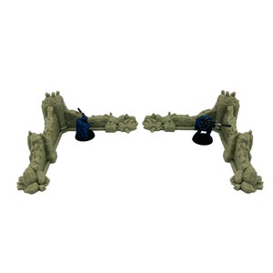 May include: Two L-shaped beige stone wall ruins for tabletop gaming, each featuring a textured, weathered surface. Two small blue-painted soldier figurines are positioned behind the walls, each holding a rifle on a black circular base.