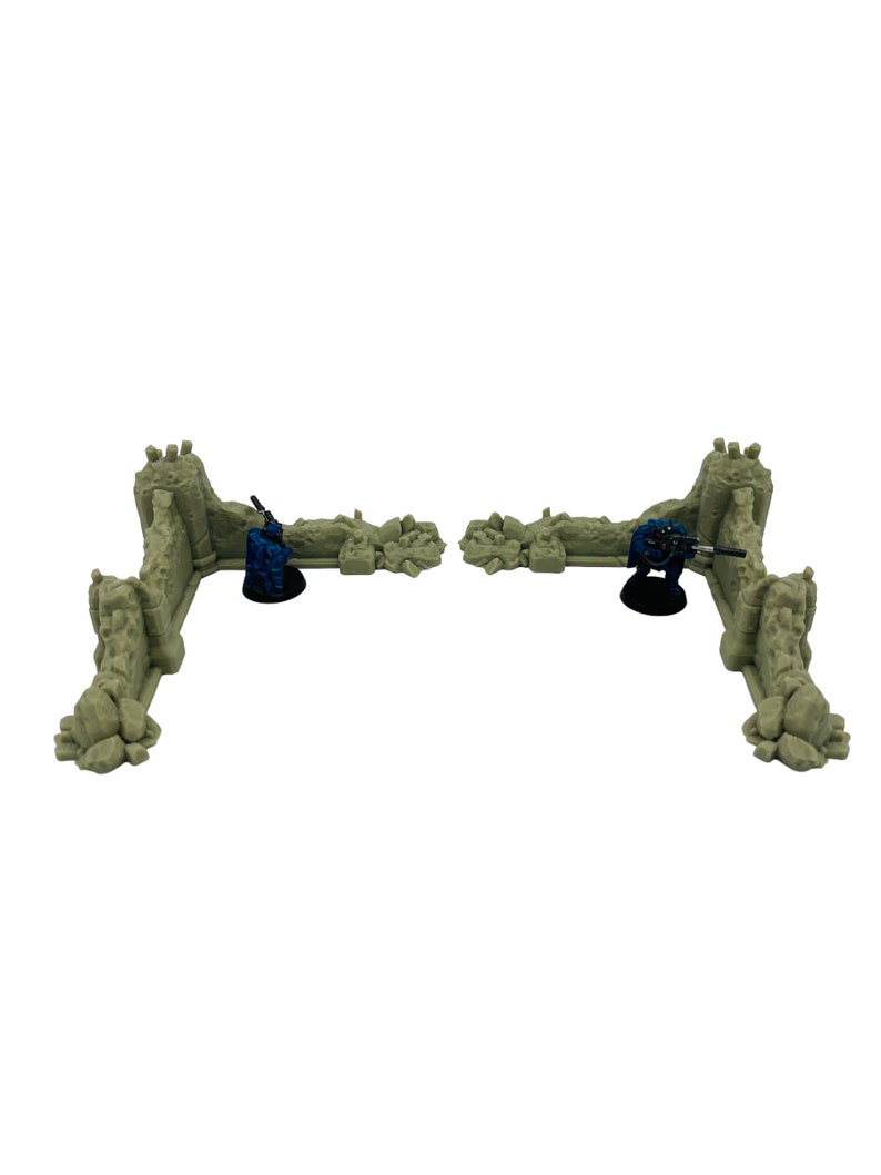 May include: Two L-shaped beige stone wall ruins for tabletop gaming, each featuring a textured, weathered surface. Two small blue-painted soldier figurines are positioned behind the walls, each holding a rifle on a black circular base.