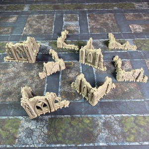 May include: A collection of eight beige, unpainted miniature building ruins featuring gothic-style arched windows and weathered stone textures. These small, modular terrain pieces are designed for tabletop gaming and display intricate architectural details.