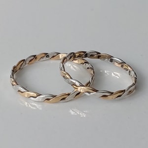 May include: Two intertwined rings with a braided design. The rings feature alternating sections of gold and silver metal, creating a textured appearance. The rings are displayed on a white surface.