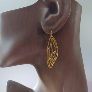 May include: Gold-toned earrings shaped like a butterfly wing. The earrings are delicate and intricate, with a detailed design.