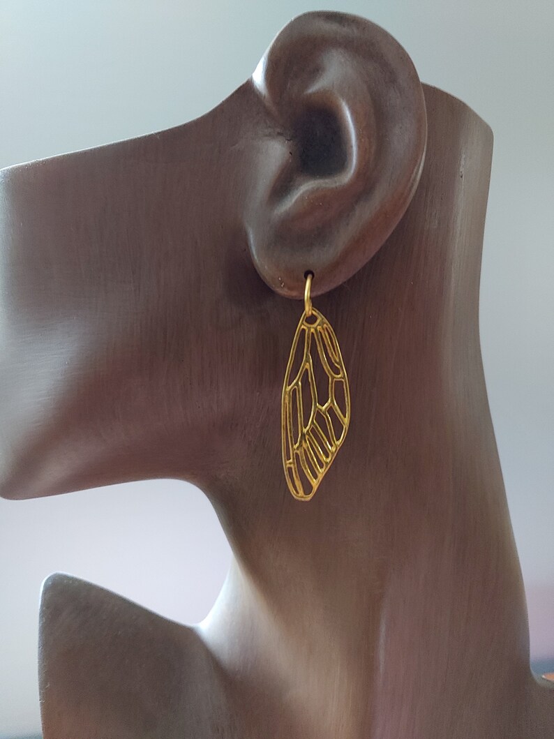 May include: Gold-toned earrings shaped like a butterfly wing. The earrings are delicate and intricate, with a detailed design.