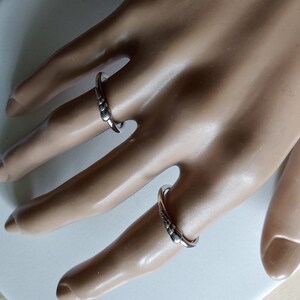 K&ouml;nnte beinhalten: Zwei silberne Ringe mit einem strukturierten Design. Die Ringe werden an den Fingern einer Mannequin-Hand getragen.